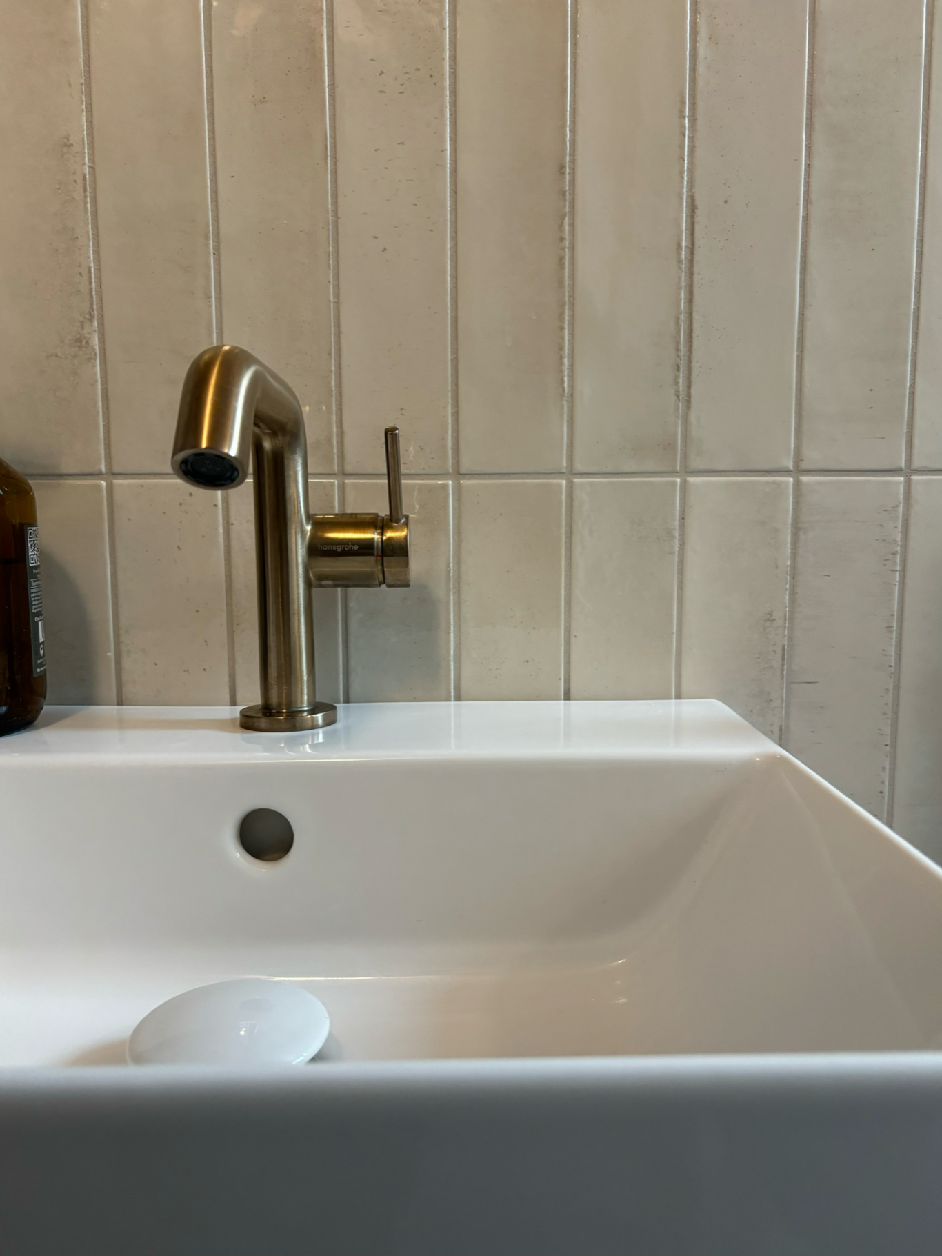Weißes Waschbecken mit bronzefarbener Armatur und Keramikfliesen im Hintergrund.