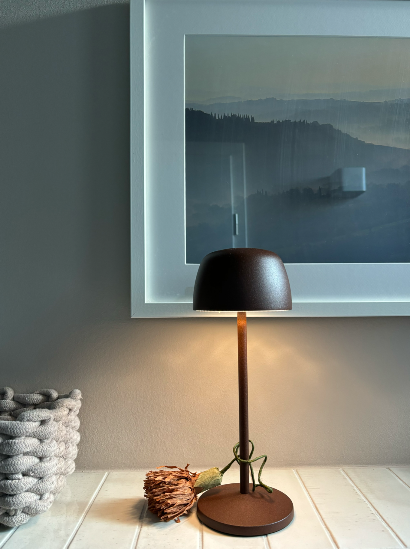 Tischlampe mit braunem Schirm, vor einem gerahmten Landschaftsbild und einer Dekokorb.